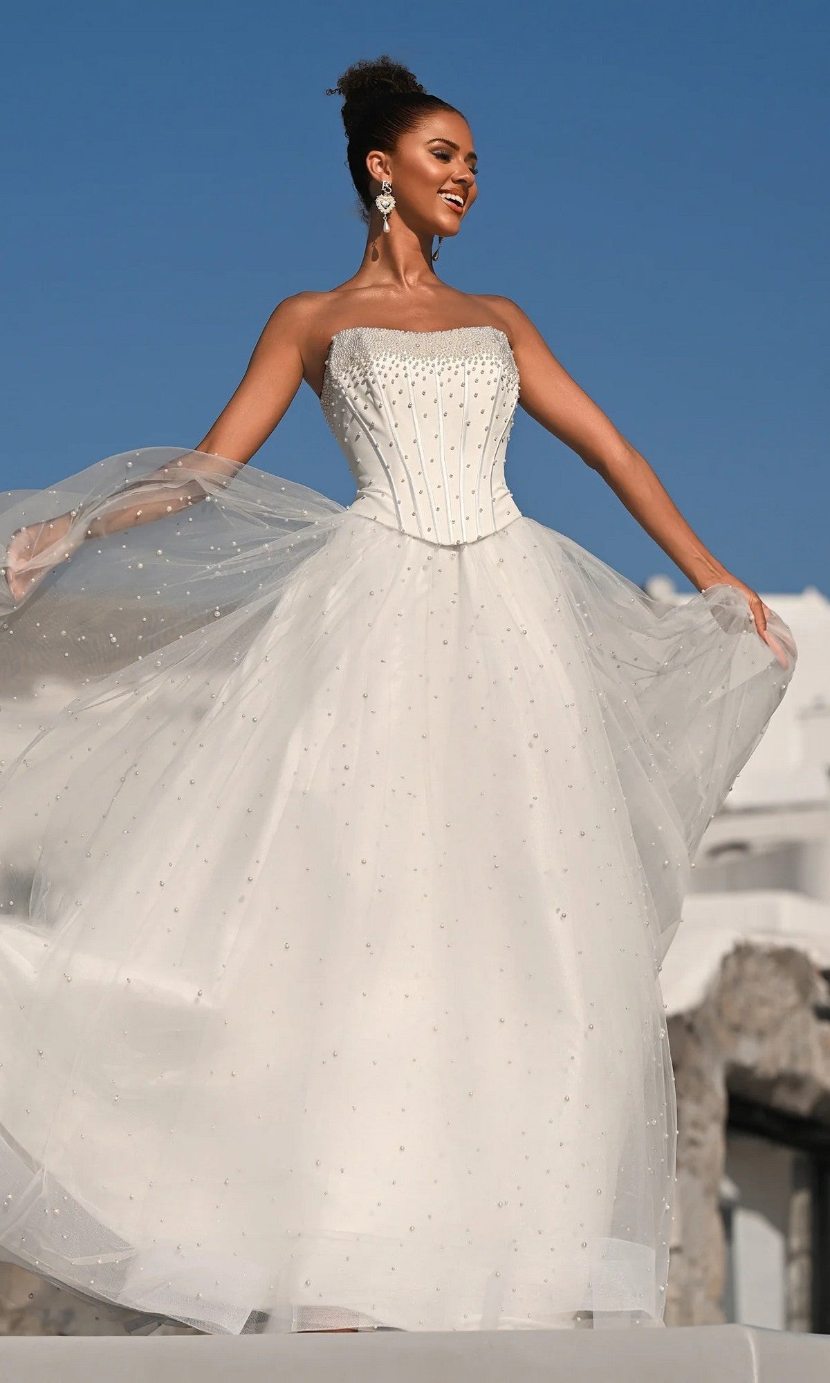 A woman stands outdoors under a clear blue sky, holding out the glitter tulle skirt of the white Portia and Scarlett PS26034 long formal dress with bead embellishments and a structured corset bodice.